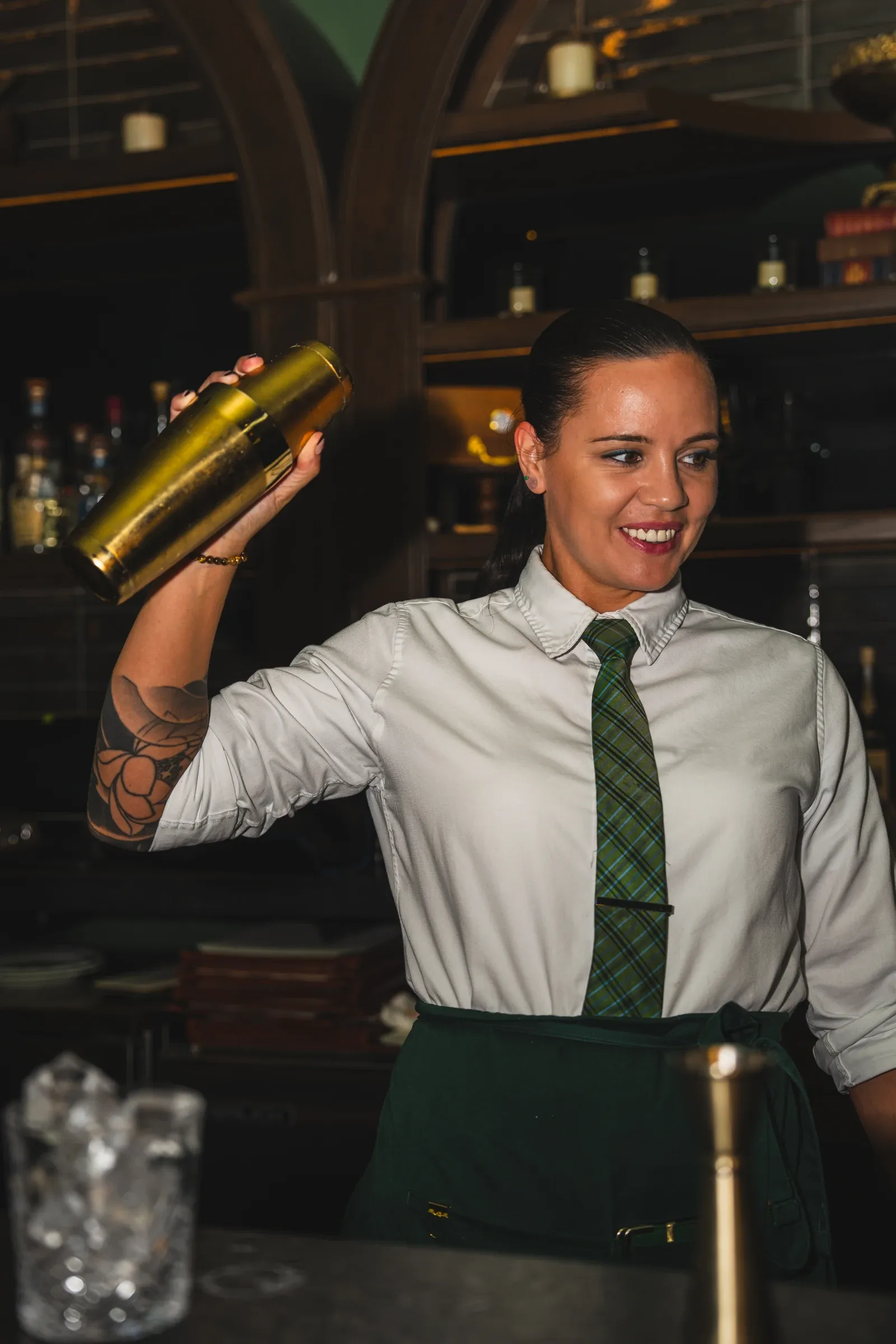 A bartender in a plaid tie shakes a gold cocktail shaker behind the bar and smiles