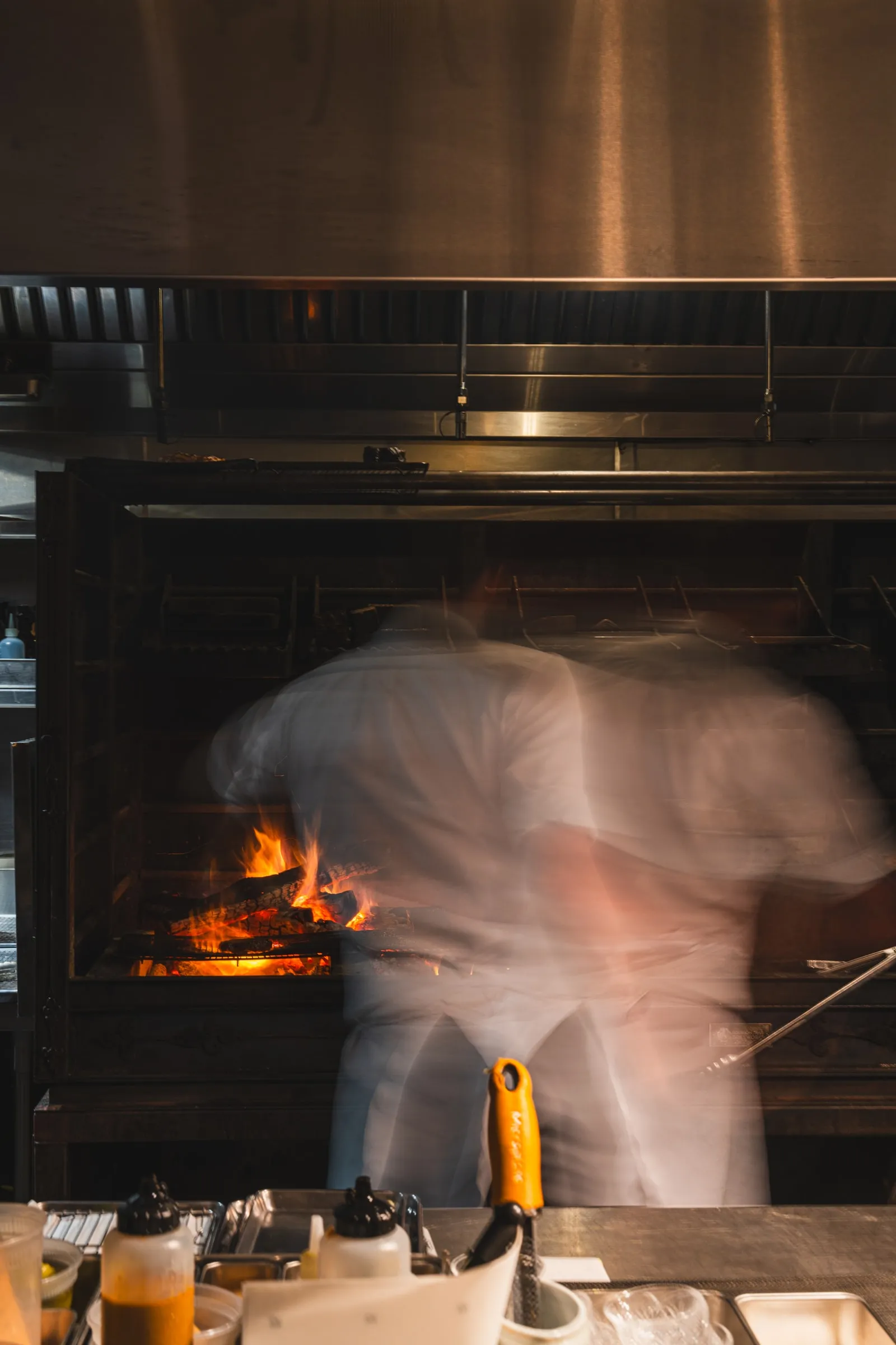 A cook at the open hearth, the fire bright behind the grill